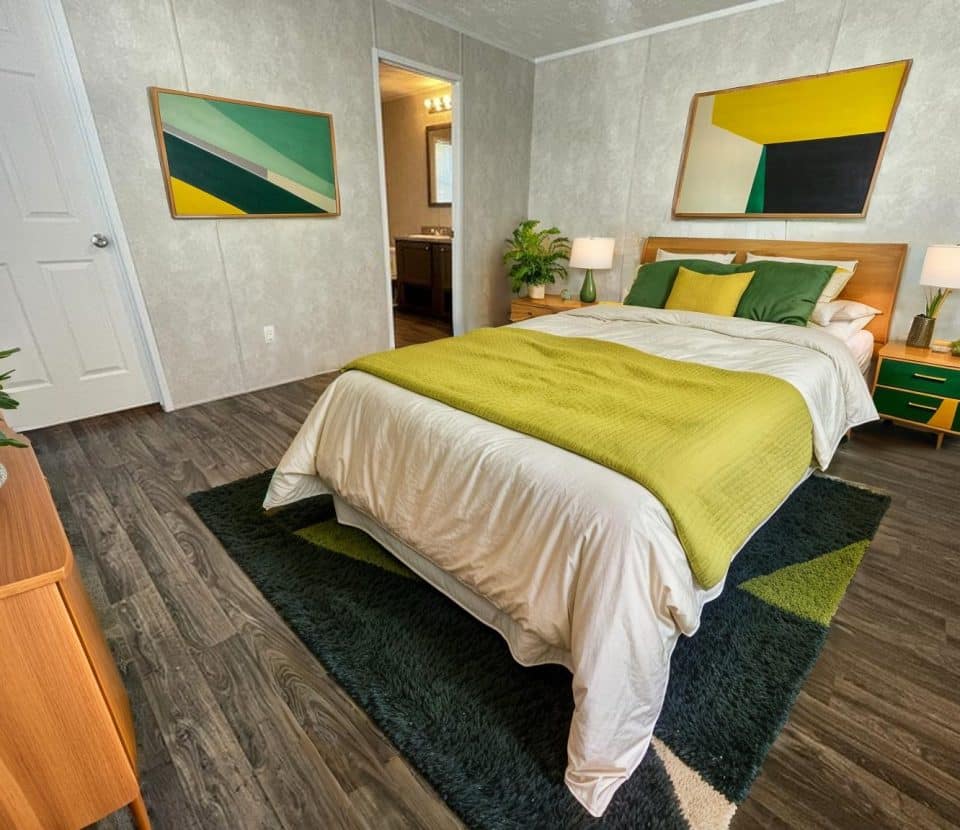 A modern bedroom with a neatly made bed featuring green and white bedding, geometric artwork on the walls, green accents, wooden furniture, and a view into a bathroom. The floor is dark wood with a green rug.