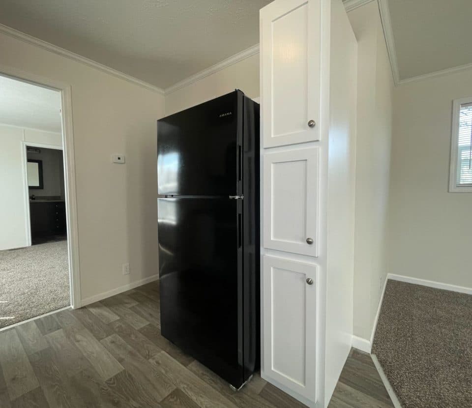 A black refrigerator stands next to white cabinets in a clean, modern kitchen with wood-look flooring and cream-colored walls. A window and carpeted adjacent room are visible in the background.