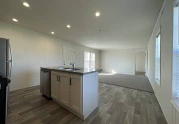 Modern kitchen with light wood island, stainless steel sink, dishwasher, and refrigerator. Open layout leads to a carpeted living area with large windows and white walls. Natural light fills the spacious room.