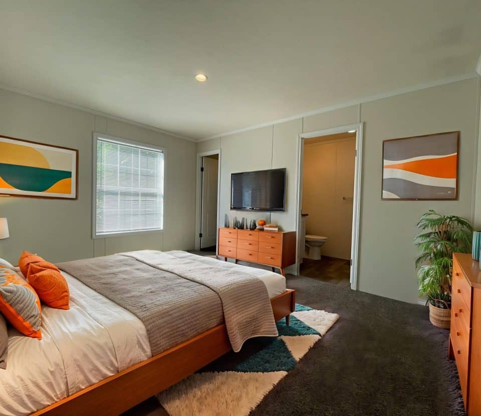 Modern bedroom with a neatly made bed, orange and gray accents, a wooden dresser, wall-mounted TV, two abstract paintings, large window, potted plant, geometric rug, and an open door leading to a bathroom.