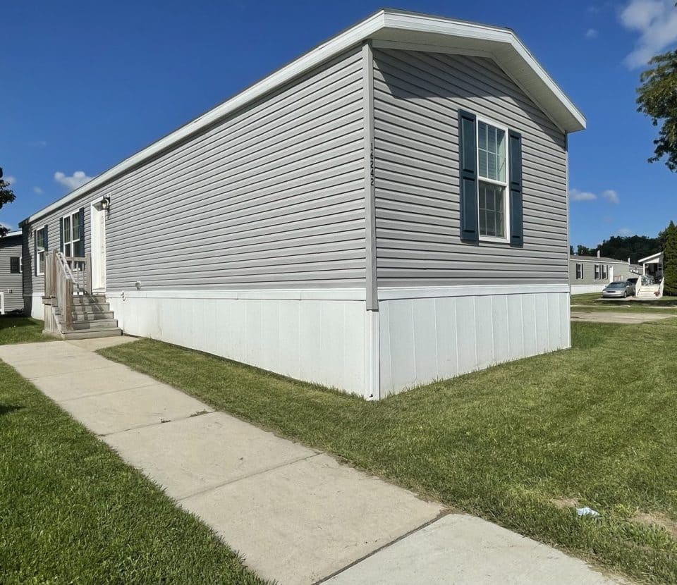 A light gray manufactured home with white trim and steps sits on a grassy lot next to a sidewalk, under a blue sky with a few clouds. Trees and other homes are visible in the background.