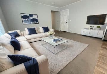A modern living room with white sofas, blue accent pillows, a glass coffee table with flowers, a wall-mounted TV, and a large rug on a neutral floor. A leaf artwork hangs on the wall above the sofa.