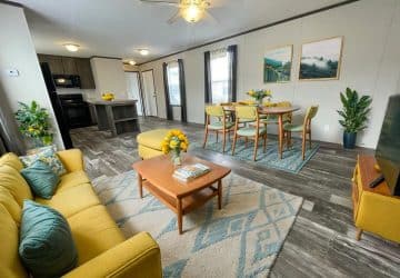 A modern, open-concept living and dining area with gray wood floors, a yellow sofa and chairs, a wooden coffee table, a dining table with six chairs, plants, and large windows letting in natural light.
