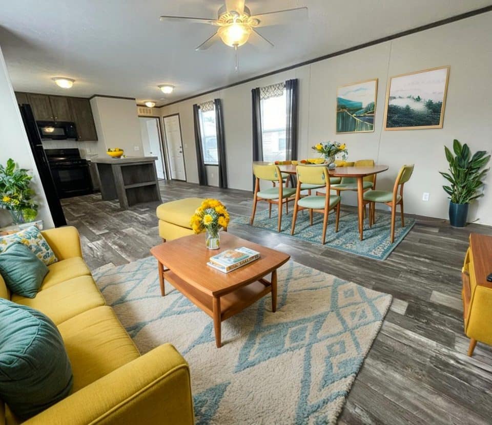 A modern, open-concept living and dining area with gray wood floors, a yellow sofa and chairs, a wooden coffee table, a dining table with six chairs, plants, and large windows letting in natural light.