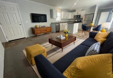 Modern living room and kitchen with gray walls, wood floors, a navy sofa with yellow pillows, a wooden coffee table, a TV on the wall, and a round dining table with chairs near a window.