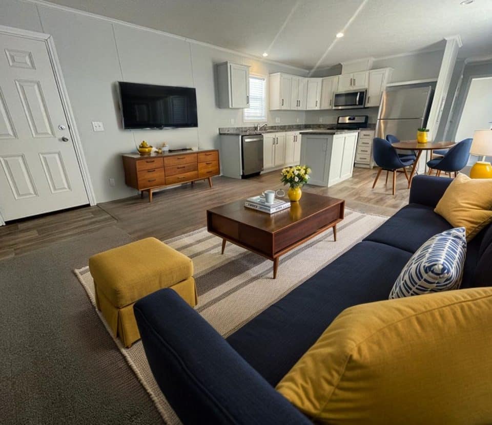 Modern living room and kitchen with gray walls, wood floors, a navy sofa with yellow pillows, a wooden coffee table, a TV on the wall, and a round dining table with chairs near a window.