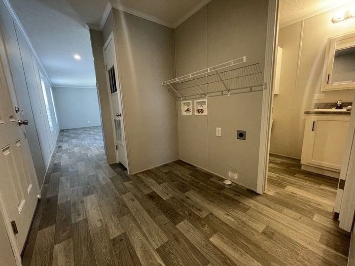 Wide-angle view of a modern, unfurnished hallway with wood-patterned flooring, beige walls, a laundry area with a wire shelf, and adjacent rooms visible, including a bathroom and living space.
