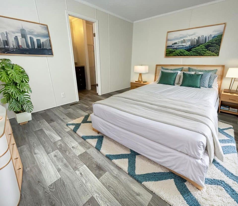 Modern bedroom with a bed, white and teal bedding, two nightstands with lamps, framed cityscape art on the walls, a potted plant, and light gray wood flooring. A doorway leads to another room.