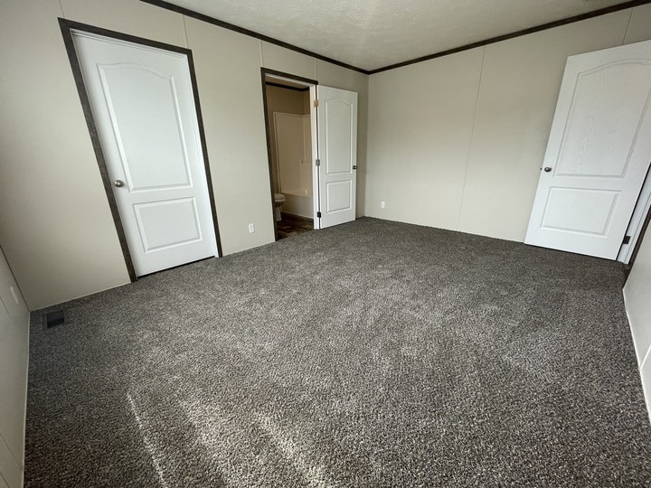 Empty room with beige walls, gray carpet, two closed white doors, and one open door revealing a bathroom. The room is well-lit with natural light coming from the left, out of frame.