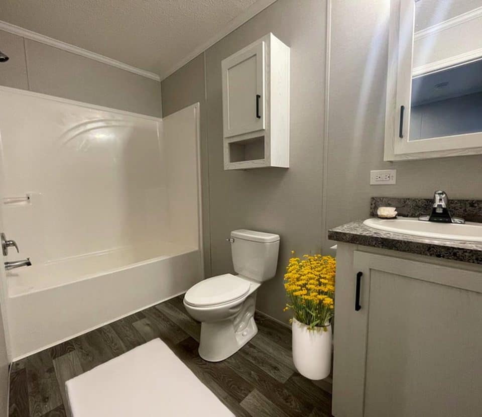 A modern bathroom with gray walls, a white bathtub-shower combo, a toilet beside a pot of yellow flowers, a gray vanity with a sink, a mirror cabinet, and wood-style flooring.