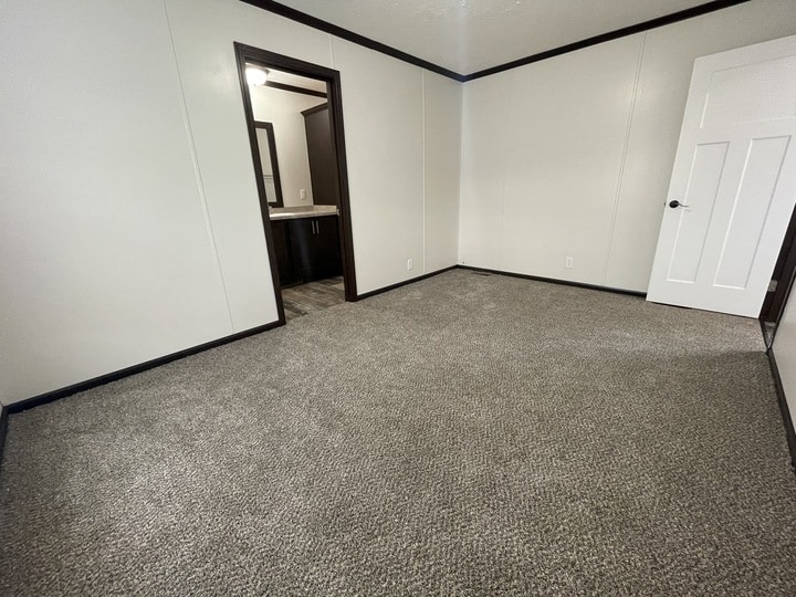 Empty room with light-colored carpet, white walls, dark trim, and a white door. An open doorway leads to a bathroom with a sink, mirror, and dark cabinets visible.