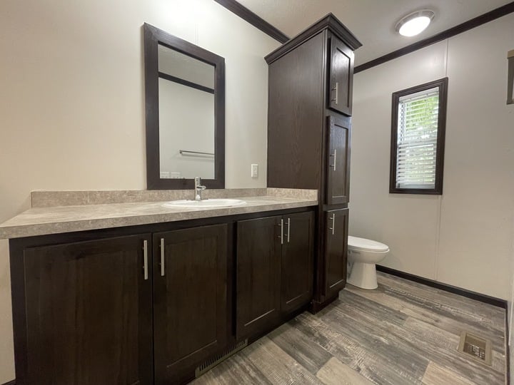 Modern bathroom with dark wood cabinets, a countertop with a sink, a mirror above, a tall storage cabinet, a toilet, and a window with blinds. The floor has gray-toned wood-like tiles.