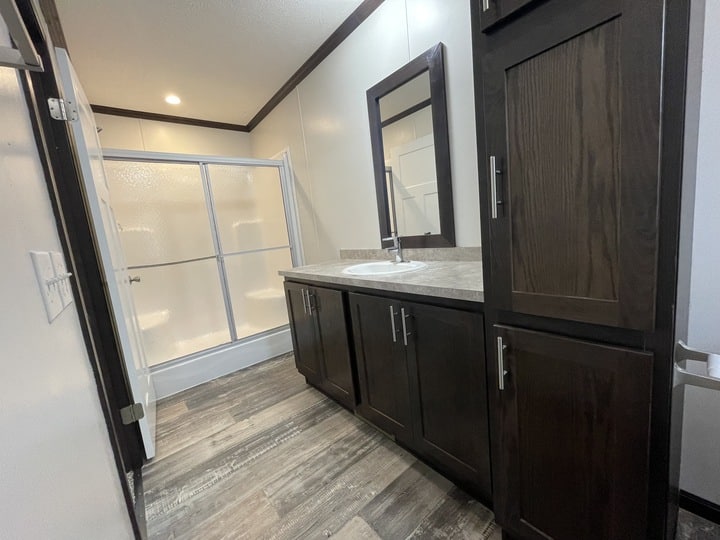 Modern bathroom with wood cabinets, a large mirror above a single sink, and a walk-in shower with sliding frosted glass doors. The floor has a wood-like finish and the walls are light-colored.