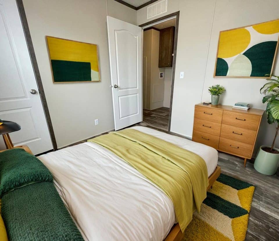 A modern bedroom with a wooden bed, green and yellow bedding, a wooden dresser, indoor plant, abstract artwork on the walls, and a door open to a hallway. The decor features green, yellow, and white accents.