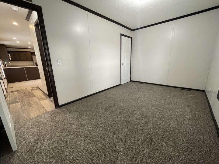 A small, empty room with beige carpet, white walls, black trim, and a closed white door. An open doorway leads to a kitchen area with wood-like flooring and visible cabinets.