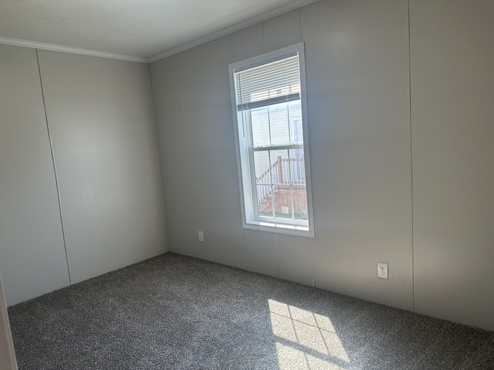 An empty room with light gray walls, a window letting in sunlight, and gray carpet flooring. The window shows a partial view of a porch and stairs outside.