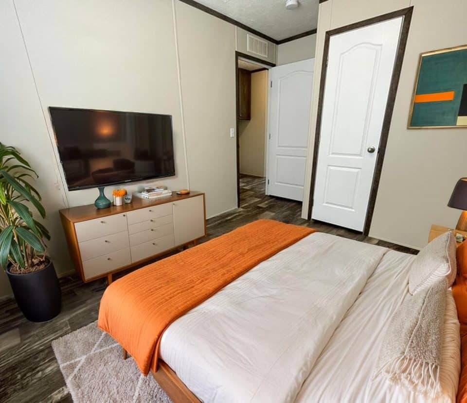 A modern bedroom with a bed featuring orange and white bedding, a TV on a wooden dresser, a potted plant, abstract wall art, and a lamp on a nightstand. The floor is dark wood, and two doors are visible.