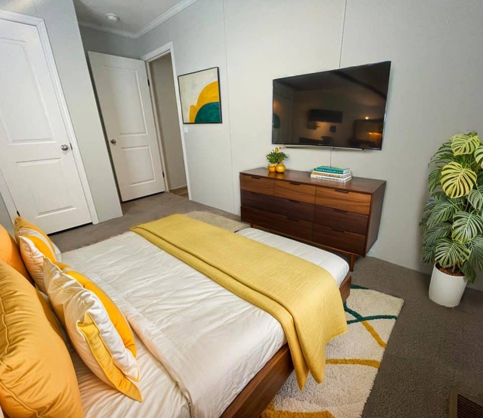 A modern bedroom with a neatly made bed featuring white and yellow bedding, a wooden dresser with a TV above it, a large green potted plant, abstract wall art, and neutral-colored walls and carpet.