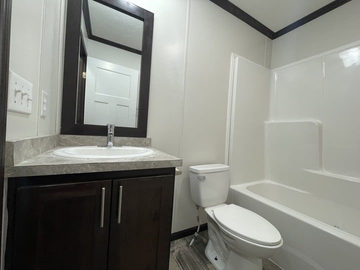 A small bathroom with a white sink and dark wood vanity, a wall mirror, a white toilet, and a white bathtub-shower combo against light-colored walls.