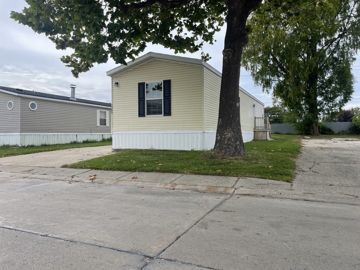 A beige mobile home with black shutters sits on a concrete lot beside a tree, with another mobile home and green lawn visible in the background under a cloudy sky.