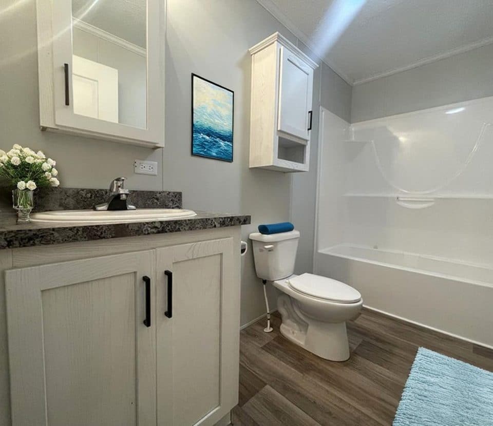 Modern bathroom with white cabinets, a dark countertop, sink with flowers in a vase, toilet with a blue cushion, bathtub, wall art, and a light blue rug on wood-style flooring.