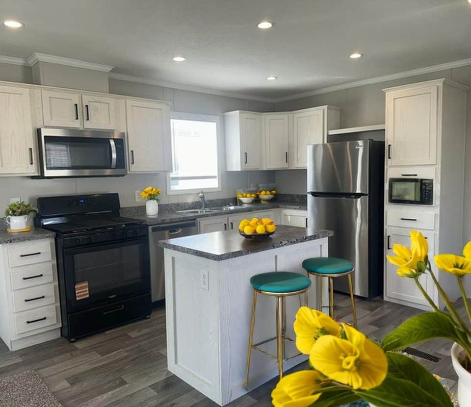 Modern kitchen with white cabinets, stainless steel appliances, an island with two teal stools, and yellow flowers and lemons as decor. Bright lighting and gray flooring create a clean, inviting atmosphere.