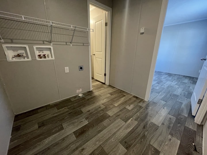 A laundry room with wood-patterned flooring, wall-mounted wire shelf, water hookups, and a door leading to a hallway and a bedroom with similar flooring.