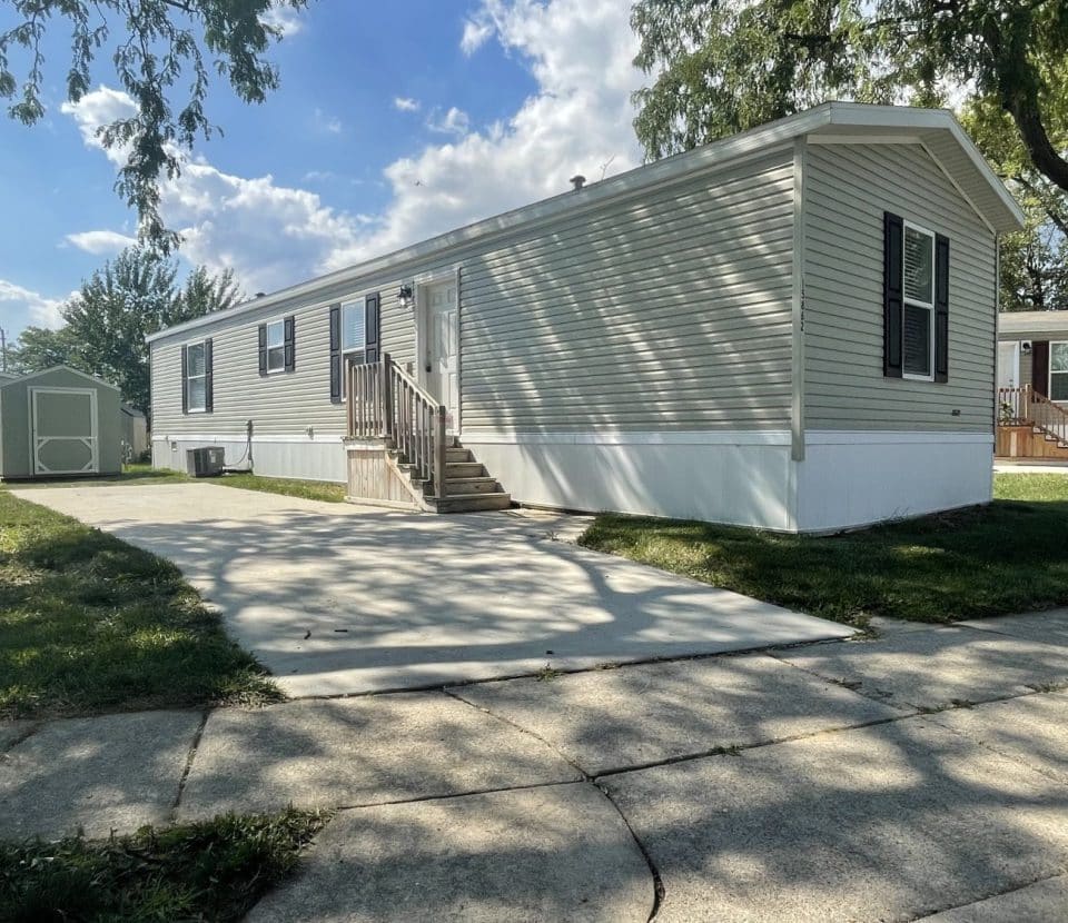 A light-colored mobile home with dark shutters, a small front porch with steps, and a driveway. Green grass, a large tree, and a small shed are visible under a blue sky with some clouds.