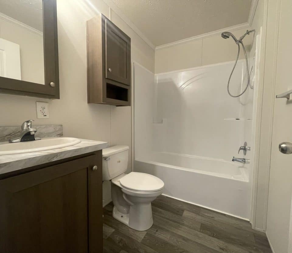 A small bathroom with a sink and dark wood vanity, a toilet, and a white bathtub-shower combo. There is a wood cabinet above the toilet and grey wood-look flooring. The walls are light-colored.