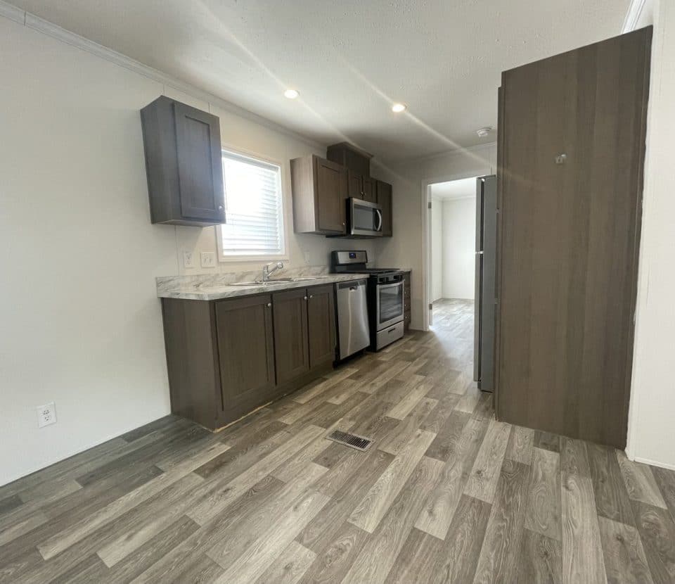 Modern kitchen with dark cabinets, marble countertops, stainless steel appliances, wood-patterned flooring, and a window above the sink. The space is well-lit and leads to another room in the background.