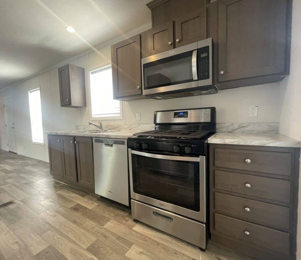 Modern kitchen with stainless steel appliances, including a stove, microwave, and dishwasher, brown cabinets, marble-look countertops, and wood-style flooring. Natural light enters through nearby windows.