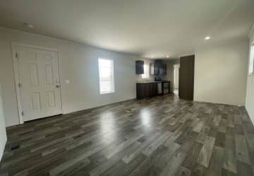 A spacious, empty room with wood-look vinyl flooring, light-colored walls, a front door, two windows, and an open kitchen area with dark cabinets and stainless steel appliances in the background.