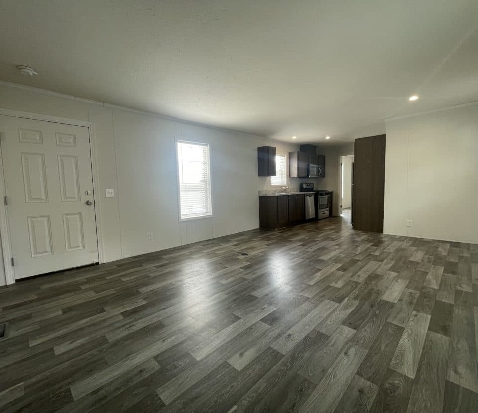 A spacious, empty room with wood-look vinyl flooring, light-colored walls, a front door, two windows, and an open kitchen area with dark cabinets and stainless steel appliances in the background.