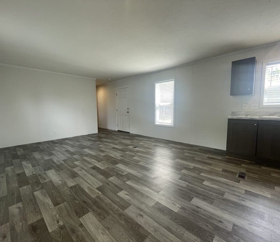Open, unfurnished room with wood-patterned flooring, white walls, a white front door, two windows, and a dark kitchen cabinet with a sink under one window. Natural light brightens the space.