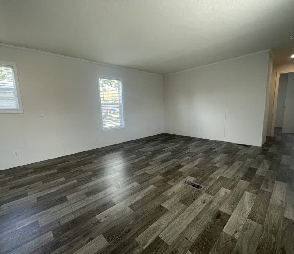 An empty room with light-colored walls, dark wood-patterned flooring, two windows letting in natural light, and a hallway leading to another area.