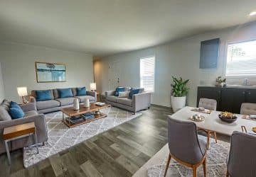 Modern living and dining area with gray sofas and blue cushions, wooden coffee tables, a dining table set for four, potted plant, and large windows letting in natural light on wood flooring.