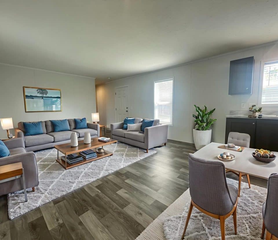 Modern living and dining area with gray sofas and blue cushions, wooden coffee tables, a dining table set for four, potted plant, and large windows letting in natural light on wood flooring.