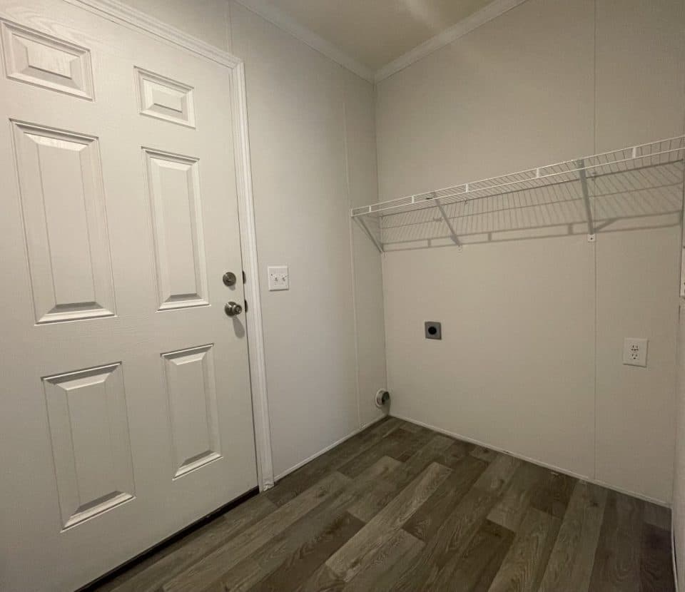 A small, empty laundry room with light-colored walls, wood-style flooring, a white door, a wire shelf, and connections for a washer and dryer.