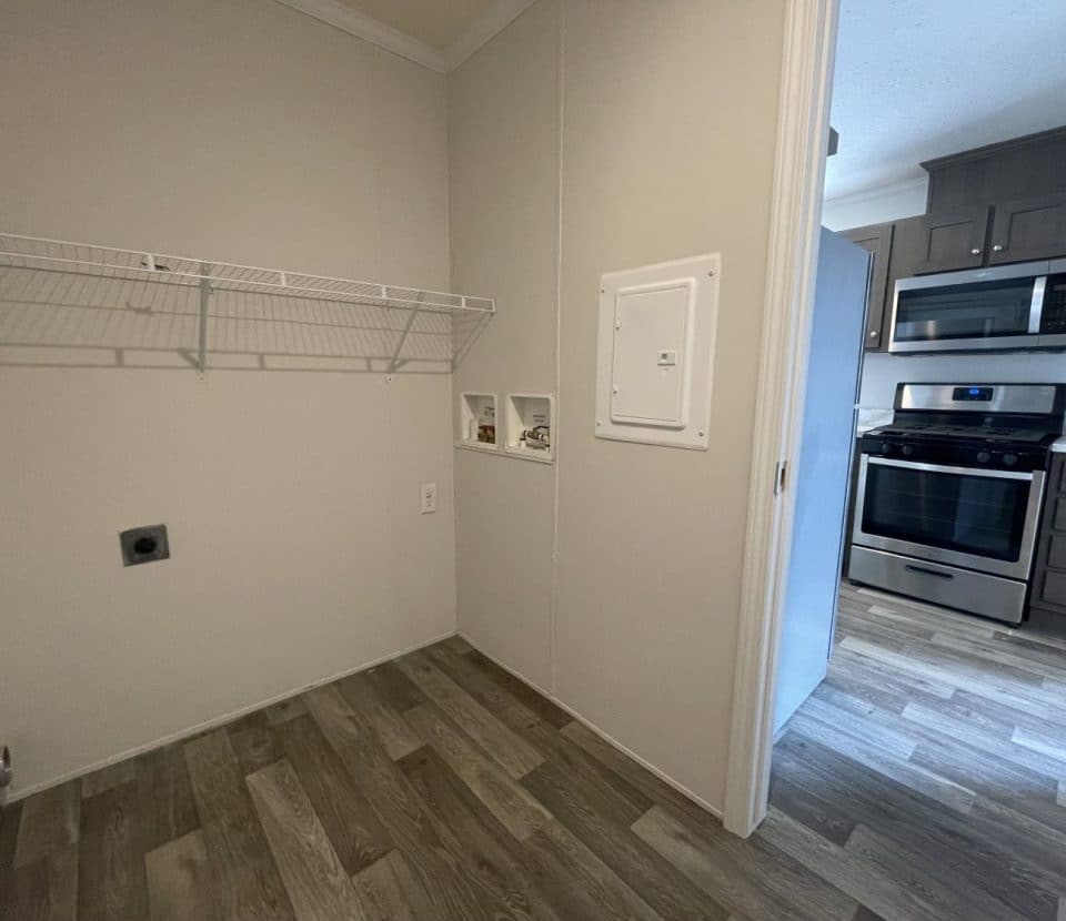 A small laundry room with a wire shelf, washer and dryer hookups, and an open doorway leading to a kitchen with stainless steel appliances and gray cabinets.