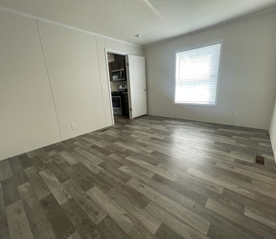 Empty room with gray wood-patterned flooring, white walls, a large window with blinds letting in natural light, and a doorway leading to a kitchen with visible appliances.