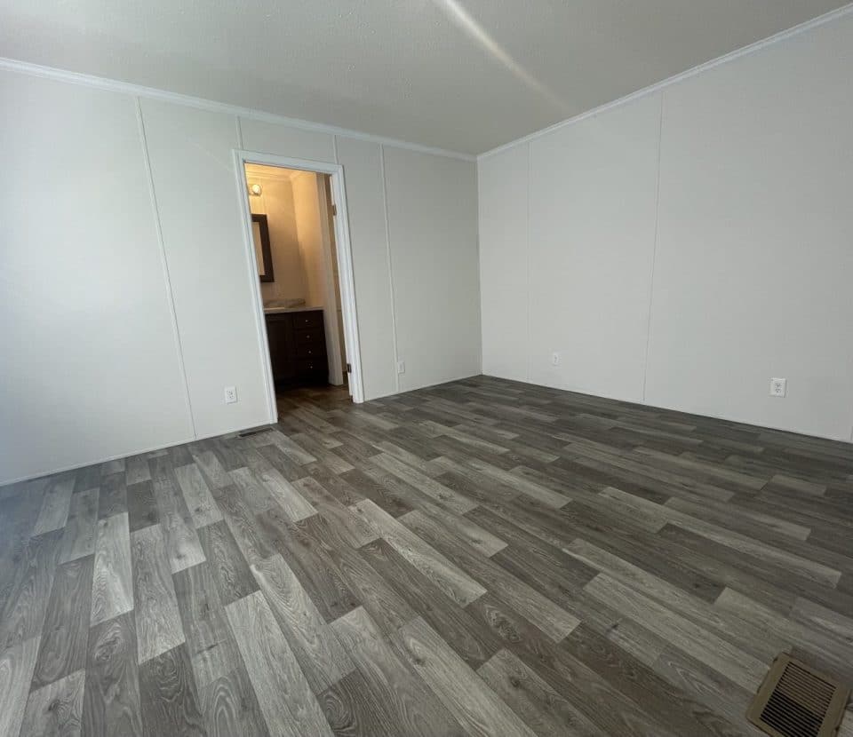 Empty room with gray wood-patterned flooring, off-white walls, and a doorway leading to a bathroom with a sink and mirror visible. Room has a vent on the floor and minimal natural light.