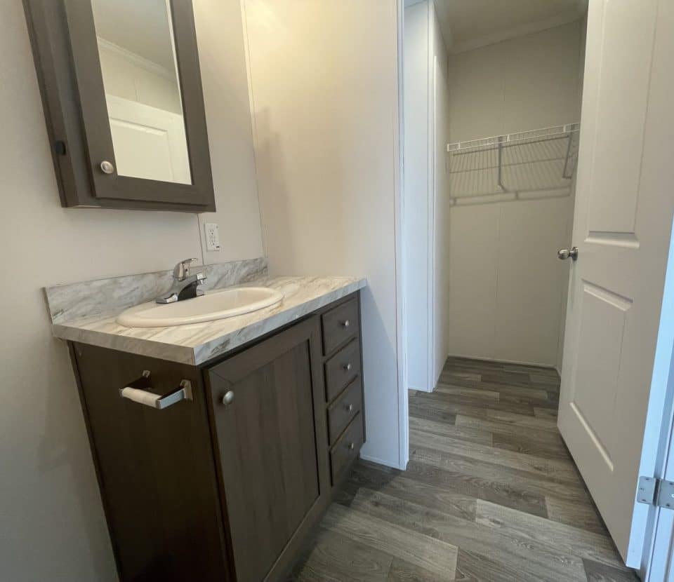 A bathroom with a dark wood vanity, marble countertop, sink, wall-mounted mirror cabinet, and a door leading to a small walk-in closet with wire shelving. The floor has wood-look vinyl planks.