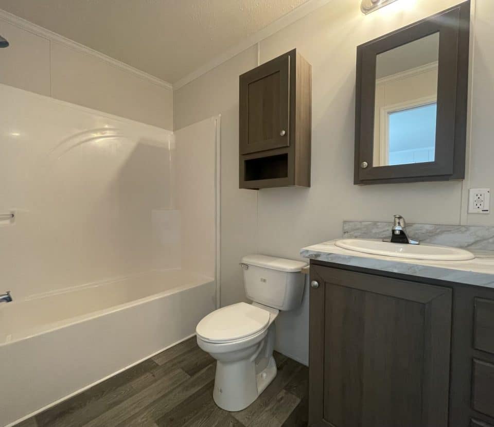 A modern bathroom with a white bathtub and shower, a toilet, a dark wood vanity with a marble countertop, a matching wall cabinet, and a mirror above the sink. The floor is gray wood-style laminate.