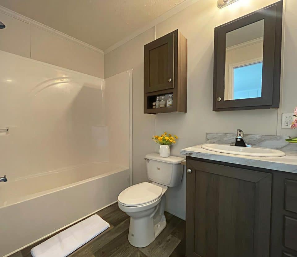 A clean, modern bathroom with a white bathtub and shower, a toilet with a yellow flower vase on top, a dark wood vanity with a sink, a mirror, and potted pink flowers on the counter. The floor is wood-style.