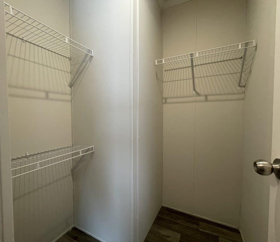 A small, empty walk-in closet with white walls, two white wire shelves on opposite sides, a wood-patterned floor, and an open door with a silver knob.