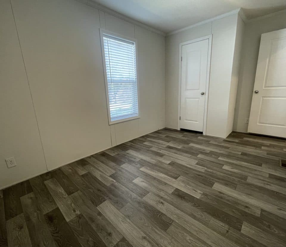 Empty room with light-colored walls, a single window with blinds, wood-patterned vinyl flooring, and two white doors; one door is slightly open, and natural light brightens the space.