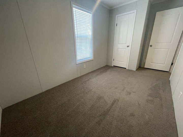 A small, empty room with light gray walls, a window with white blinds, brown carpeted floor, and two closed white doors. Natural light enters through the window.