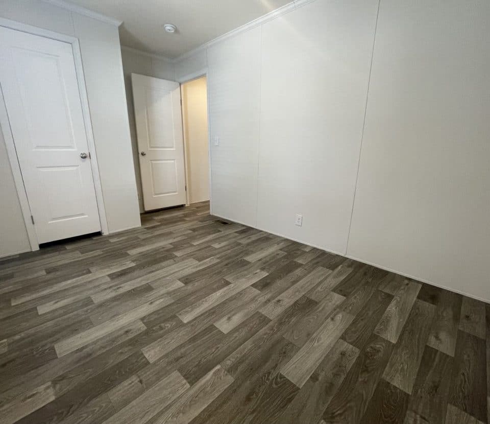 Empty room with light-colored walls, dark wood-patterned vinyl flooring, two white doors, and baseboards, lit by natural light. The overall atmosphere is clean and minimalistic.