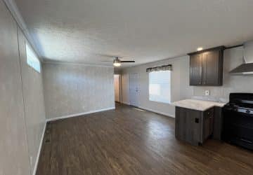 A modern, unfurnished living room with wood flooring, beige walls, ceiling fan, and an open kitchen area with dark cabinets and a black stove. Natural light enters from windows on the right.