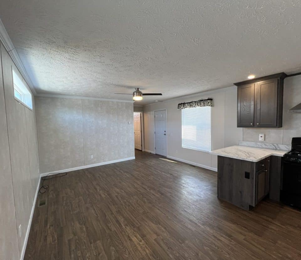 A modern, unfurnished living room with wood flooring, beige walls, ceiling fan, and an open kitchen area with dark cabinets and a black stove. Natural light enters from windows on the right.